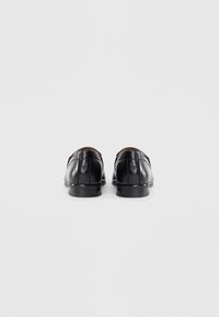 Une paire de mocassins en cuir noir vue de l'arrière, montrant des logos en relief au-dessus des talons sur un fond blanc uni.