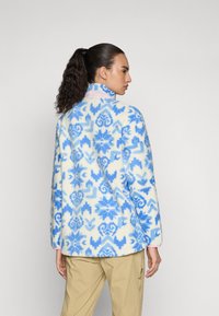 Chaqueta de forro polar con un patrón floral en azul y blanco, cuello alto y corte relajado. La textura de la tela parece suave y cálida.