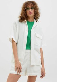 Camisa de botones blanca texturizada con un bolsillo, combinada con una blusa verde ajustada y pantalones cortos blancos a juego. La modelo lleva gafas de sol con tintes verdes.