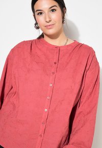 Camicia a maniche lunghe, ampia e di colore corallo, con un motivo floreale texturizzato, scollatura rotonda e bottoni frontali in tonalità abbinate.