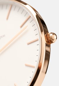 Närbild av en klocka i guldton med en vit urtavla, minimala timmarkörer och en räfflad krona på höger sida.