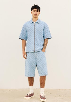 Jeune homme debout, une main dans la poche, portant une veste zippée à manches courtes à carreaux bleu clair et un short assorti longueur genou avec des baskets.