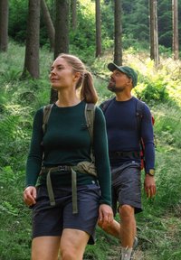 Två vandrare på en skogsstig: den ena i en mörkgrön långärmad tröja och shorts, den andra i en marinblå tröja och grå shorts, båda med ryggsäckar.