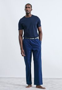 Mornarinski pamukasti majica u kombinaciji s mornarskim hlačama na crte, s crno-bijelim brendiranim pojasom. Model stoji bosa na svijetlosivoj površini.