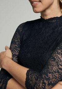 Marinefarbene Spitzenoberteil mit langen Ärmeln, hohem Kragen und filigranen Blumenmustern; weicher, strukturierter Stoff mit gewellten Kanten.
