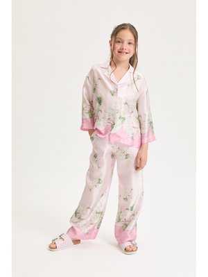 Fată zâmbitoare, stând cu mâinile în buzunare, purtând pijamale din satin roz cu flori și papuci strălucitori pe un fundal alb.