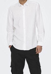 Camisa blanca de lino de manga larga con cuello abotonado y cierre frontal de botones, combinada con pantalones cargo negros. Textura suave, ajuste relajado.