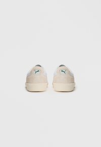 Hvite og beige nubuck Puma-sneakers vist fra baksiden, plassert ved siden av hverandre på en enkel hvit bakgrunn.
