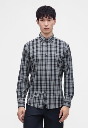 Jeune homme portant une chemise à carreaux grise et noire à manches longues, debout devant un fond clair uni.
