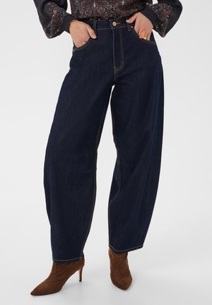 Frau trägt weite dunkelblaue Jeans mit braunen High-Heel-Stiefeln und einer gemusterten langärmeligen Bluse, Hände in den Taschen.