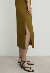 Robe vert olive avec une fente latérale, en tissu lisse, associée à des sandales noires à brides, à semelles plates et au design minimaliste.