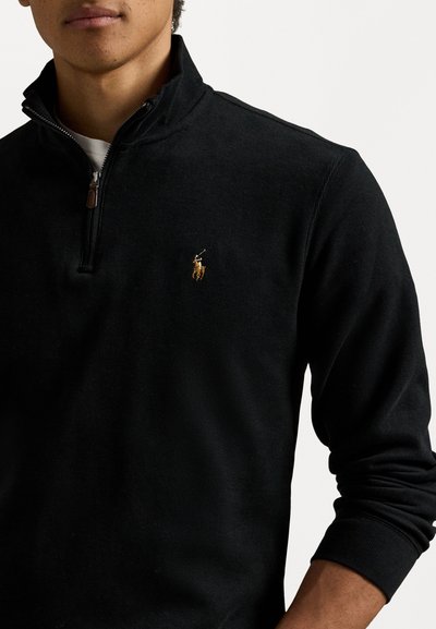 Άνδρας φορεί μαύρο pullover με κουμπί και μικρό κεντημένο λογότυπο ποδοσφαιριστή στο στήθος, στέκεται μπροστά σε λευκό φόντο.