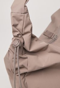 Beige zachte stoffen tas met een gedraaid handvat, voorzien van een schuifsluiting en een gestructureerd oppervlak. Het ontwerp legt de nadruk op plooien en gladde hardware.