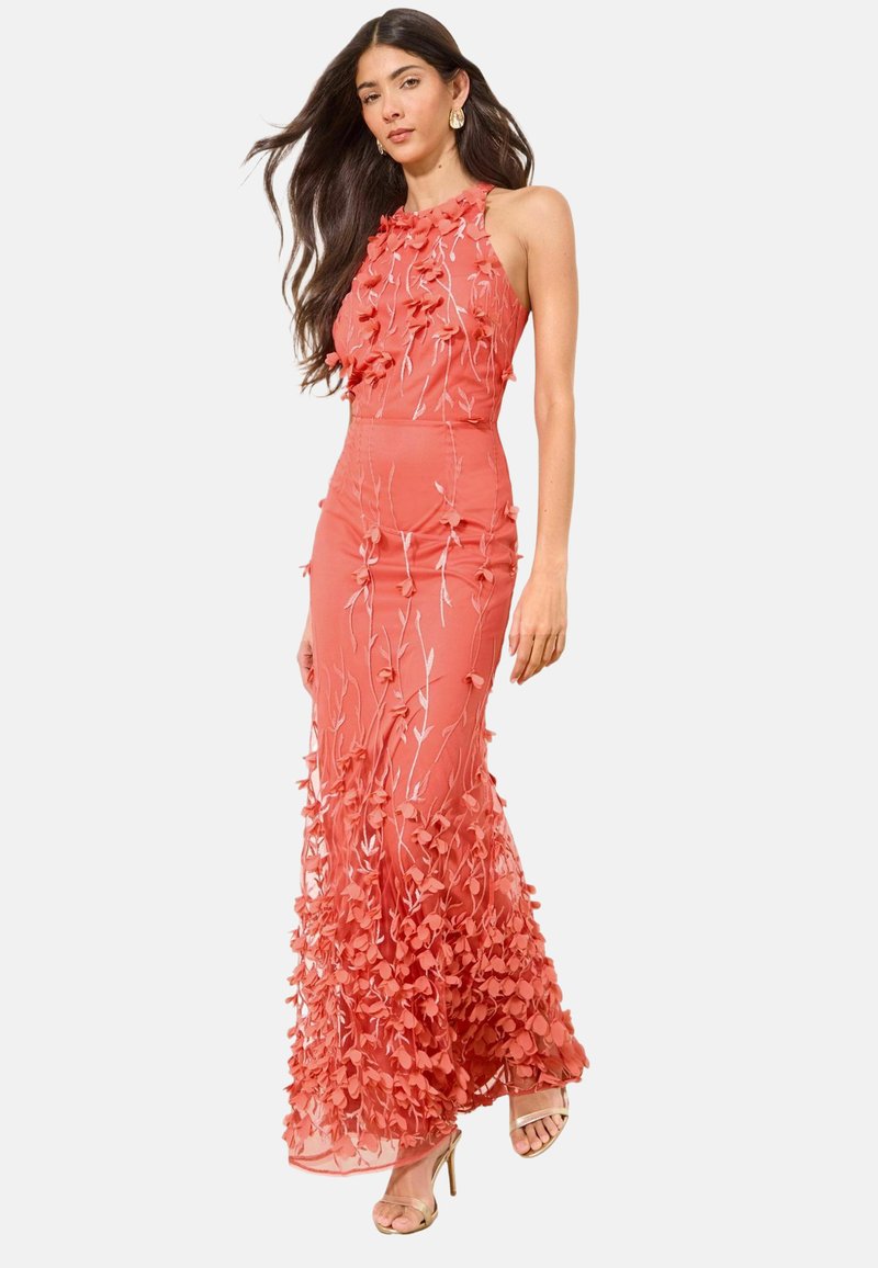Femme portant une robe maxi corail rose à fleurs sans manches avec broderie 3D, col haut et sandales beiges à talons hauts.
