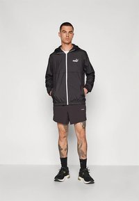 Mladić stoji u crnoj Puma jakni, crnim šortsevima, crnim patikama i crnim čarapama, pokazujući tetovaže na svojim nogama pred jednostavnom pozadinom.