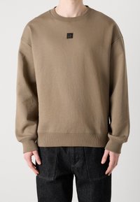 Persona che indossa una felpa beige a maniche lunghe, comoda e con polsini e orlo a coste, abbinata a jeans in denim scuro.
