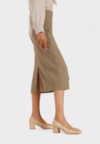 Scarpe con tacco a blocco beige con dettaglio a fibbia quadrata, abbinate a una gonna verde oliva al ginocchio con uno spacco laterale. Texture liscia.