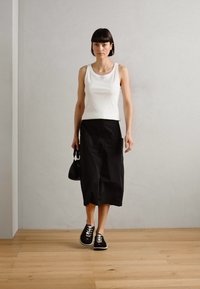White sleeveless top, black midi skirt, black sneakers, and small handbag. The floor is wooden, and the wall is light gray with a smooth texture.