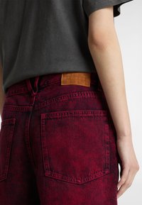 Los jeans de mezclilla rojos presentan un parche de cuero texturizado en tono marrón en la parte posterior, con costuras visibles y bolsillos traseros, combinados con una camisa gris.