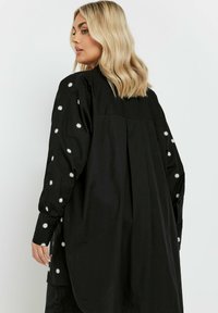 Maglietta nera a maniche lunghe, con ricamo di margherite bianche sulle maniche, vestibilità ampia e design con pieghe sul retro.