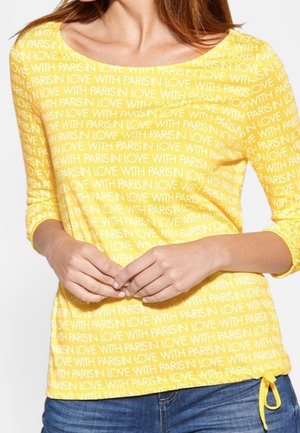 Kobieta w żółtej koszuli z długim rękawem z powtarzającym się białym napisem "LOVE WITH PARIS" oraz w niebieskich dżinsach, z rękami delikatnie złożonymi na przodzie.