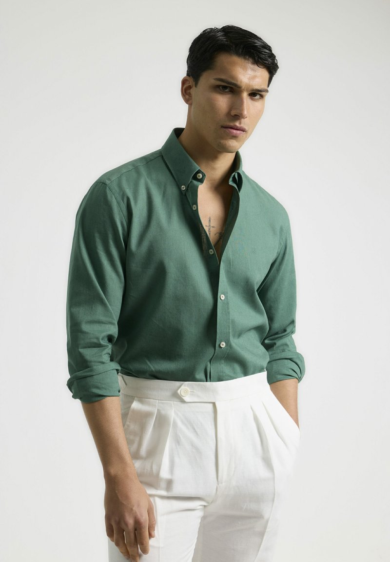 Camisa verde de botones en tejido ligero, con collar y mangas arremangadas, combinada con pantalones blancos con pliegues.