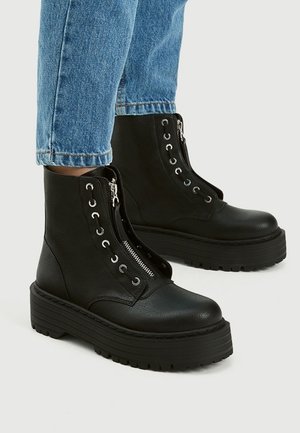 Bottes compensées en cuir noir à la cheville avec fermeture éclair sur le devant et œillets pour lacets, portées avec un jean en denim bleu.