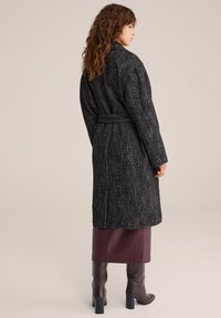 Dolga črno-bela plašča v ribjo kost s pasom, z ovratnikom z zarezo in spuščenimi rameni, kombinirana s škrtim škornjem v burgundski barvi in rjavimi čevlji.