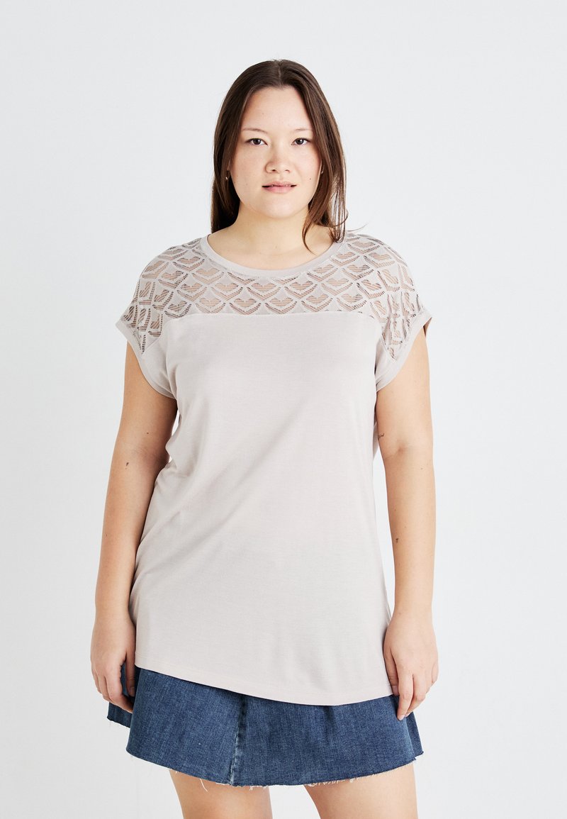 Hellbeige T-Shirt mit einer strukturierten Spitzenüberlage an den Schultern. Kurze Ärmel und lockere Passform, kombiniert mit einem Jeansrock.