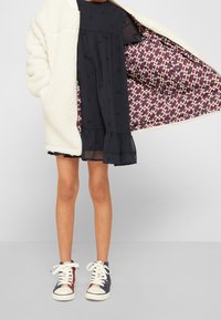 Krémová fleecová bunda s vzorovaným červeným a námořnickým vnitřkem, nošená přes černé volánkové šaty a modré, červené a bílé vysoké tenisky.