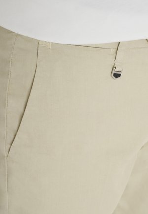 Pantalones beige a medida con bolsillos frontales y un pequeño cierre decorativo de metal plateado cerca de la cintura, combinados con una blusa blanca.