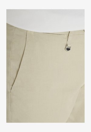Pantalones beige a medida con bolsillos frontales y un pequeño cierre decorativo de metal plateado cerca de la cintura, combinados con una blusa blanca.