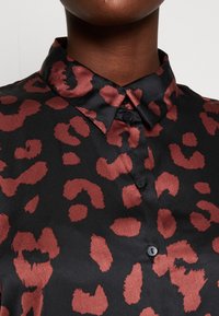 Chemise en satin noir avec un motif léopard en rouge profond. Dotée d'un col classique et d'une fermeture à boutons, mettant en avant la texture et le design.