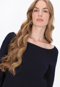 Pulover bleu marin cu umeri descoperiți, dintr-un material moale și tricotat. Prezintă o croială lejeră și un model texturat, completat de părul ondulat și desfăcut.