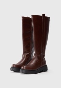 Botas de cuero marrón hasta la rodilla con una superficie lisa, punta redondeada y suela gruesa de goma negra; cuenta con un detalle de costura mínima y una lengüeta en la parte trasera.