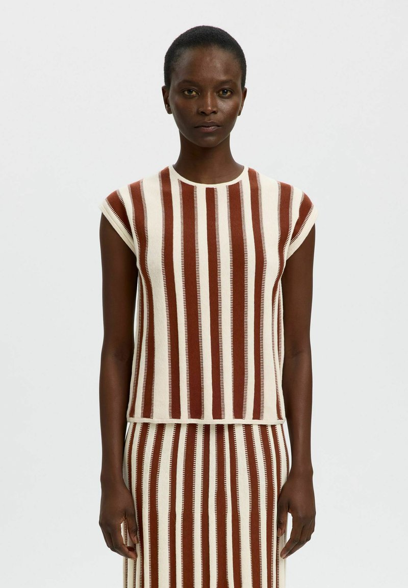 Femme portant un haut sans manches rayé verticalement marron et blanc et une jupe assortie, debout devant un fond uni.