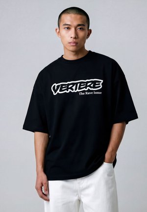 Hombre con el cabello corto, vistiendo una camiseta negra de gran tamaño con el texto "VERTIERE The Rave Issue", combinado con pantalones blancos, posando frente a un fondo gris.