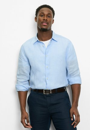 Mann mit kurzen, lockigen Haaren, trägt ein hellblaues, geknöpftes Hemd über einem weißen T-Shirt und dunkle Hosen, steht vor einem weißen Hintergrund.