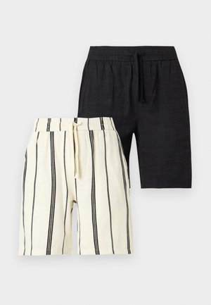 Mann mit geflochtenem Haar und Sonnenbrille, der ein schwarzes „New York“-T-Shirt, schwarze Shorts, weiße Socken und schwarze Loafers trägt und mit einer Hand in der Hosentasche steht.