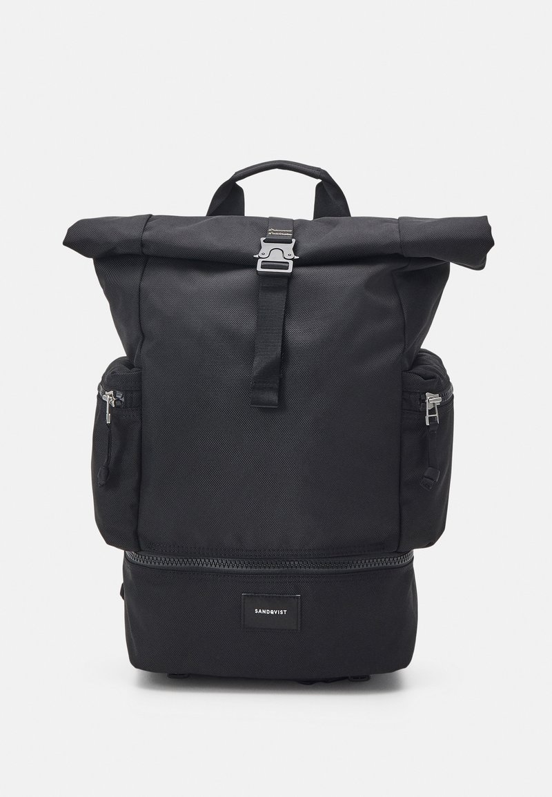 Sac à dos noir à rabat fabriqué en tissu durable, comportant deux poches zippées sur les côtés, une fermeture éclair à l'avant et un patch logo en bas.
