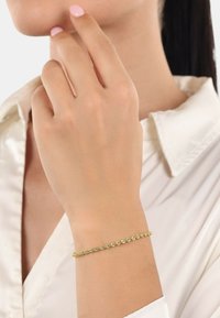 Pulsera trenzada de oro en una muñeca; textura suave, diseño delgado, combinado con un tono de piel claro. Llevada con una camisa blanca de cuello.