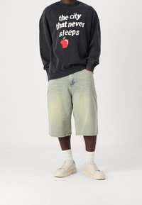 Sudadera negra deslavada con texto blanco y gráfico de una manzana, combinada con pantalones cortos de mezclilla claros, calcetines blancos y zapatillas beige.