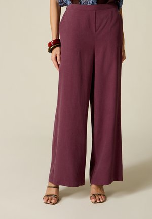 Vrouw draagt hooggetailleerde, wijd uitlopende pruimkleurige broek met twee armbanden aan de linkerpols en open sandalen met dunne bandjes.