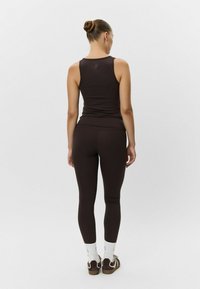 Hnedý priliehavý tielko a legíny vyrobené z hladkého materiálu, s jemným logom. Model má na sebe hnedé tenisky a biele ponožky.