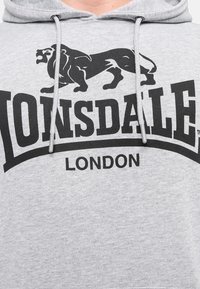 Siva dukserica s grafikom crnog lava i natpisom "LONSDALE LONDON". Izrađena od pamuka, s kapuljačom i mekanom teksturom.