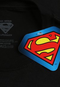 Black cotton t-shirt with a round neckline. Features a blue price tag with a red and yellow "Superman" logo prominently displayed.