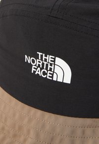 Zwarte en bruine bucket hat van stof, met een wit logo "The North Face" op de voorkant, en opvallende stiksels op de rand.