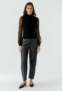 Femme se tenant debout dans un pull à col roulé noir avec des manches en dentelle transparente, un pantalon en cuir noir et des chaussures à talons aiguilles noires sur fond blanc.