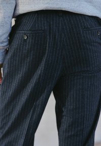 Tumši svītru bikses, izgatavotas no teksturēta auduma, ar divām aizmugurējām kabatām ar pogu aizdari, pielāgota piegriezuma un strukturēta dizaina.