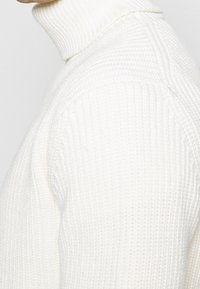 Weißer, gerippter Rollkragenpullover mit strukturiertem Strickmuster und taillierter Silhouette. Der Kragen ist hoch und enganliegend, was Wärme und Komfort betont.